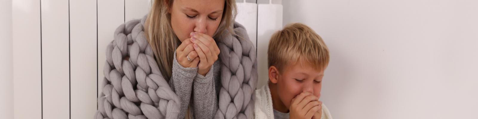 Mother and son huddle under warm blankets against a white paneled wall as they blow into their hands to stay warm.