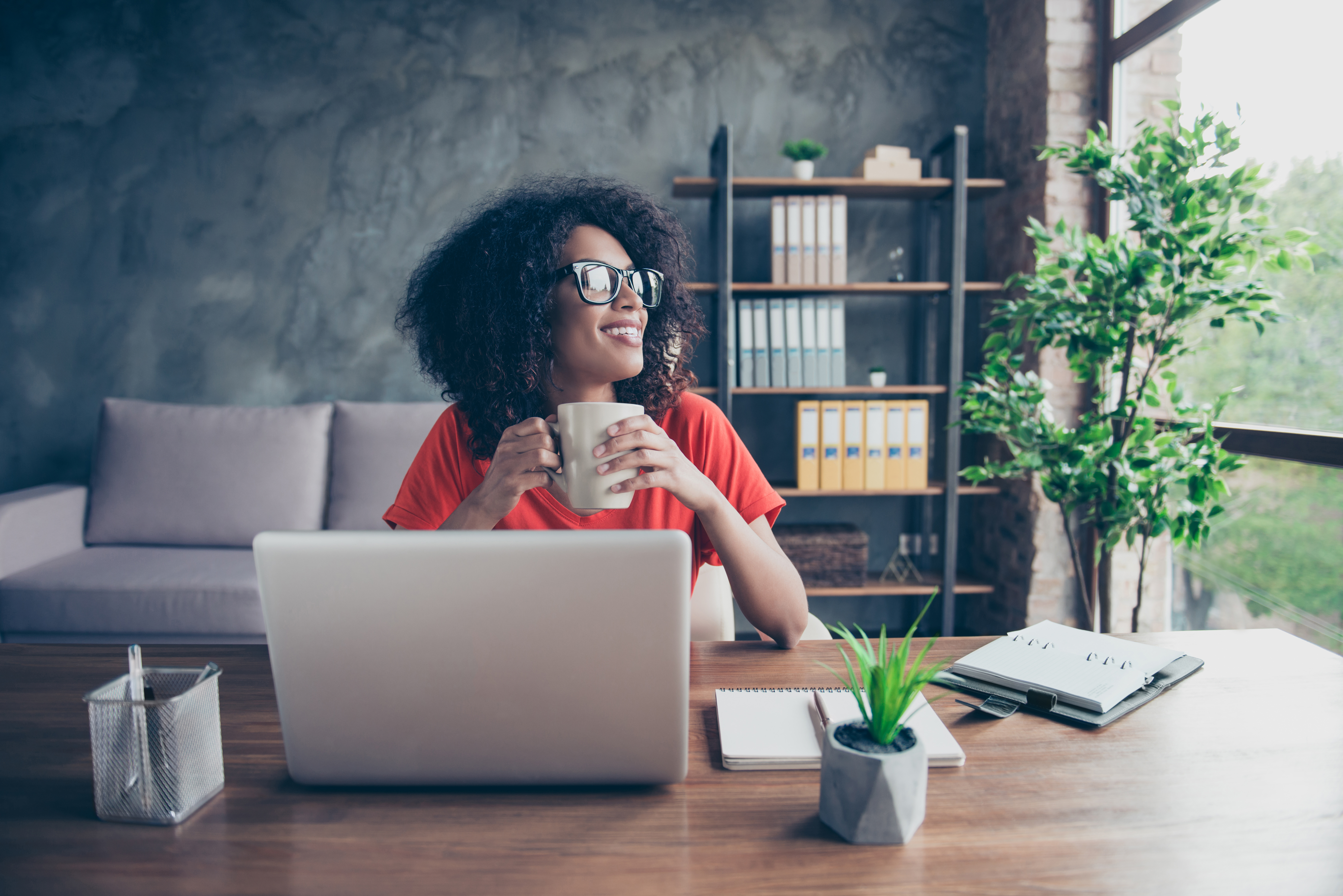 Portrait of dreamy lovely accountant sitting at desk in modern office with interior drinking hot beverage holding cup with tea in hands looking at window enjoying freetime