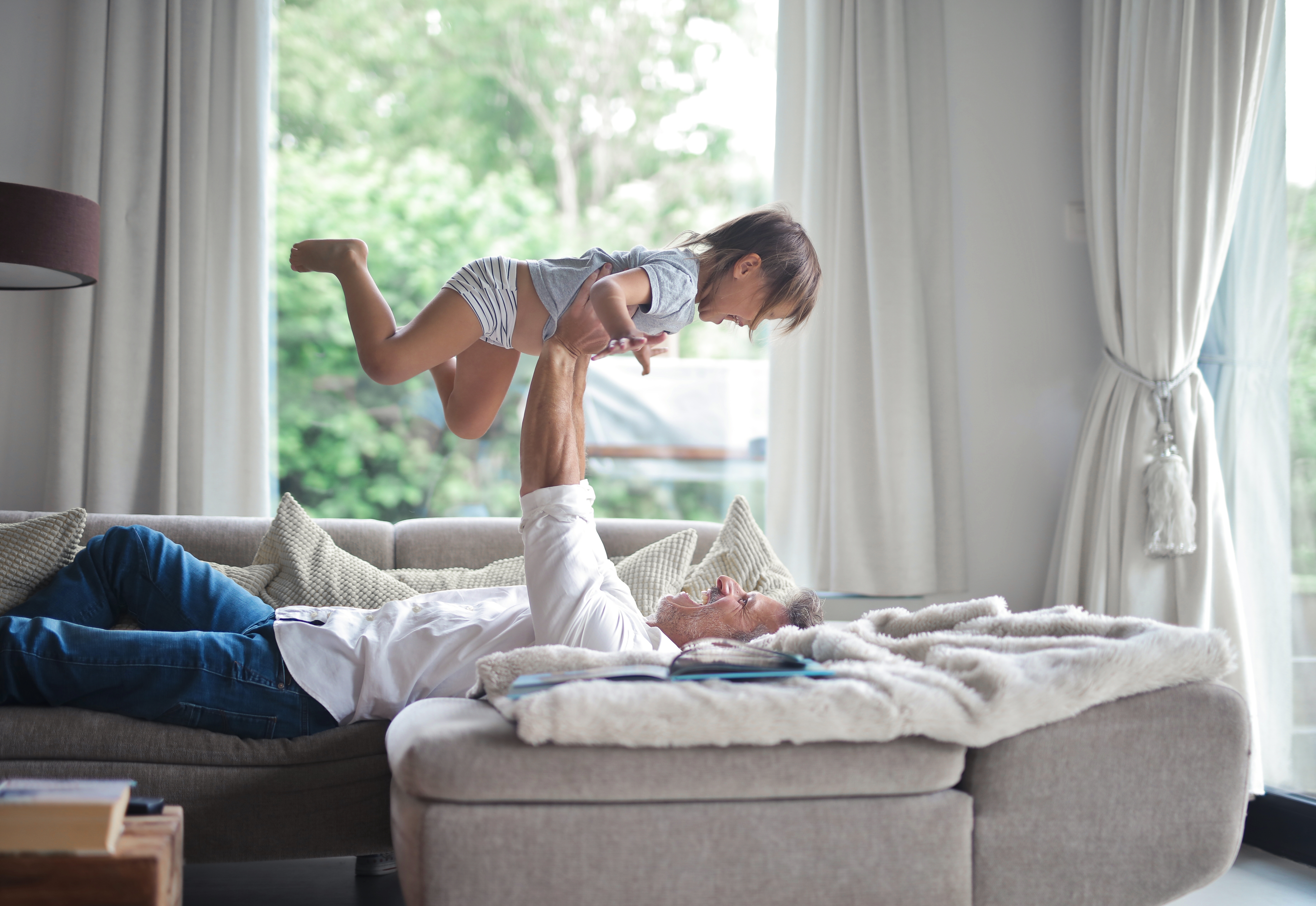 Dad lying on the sofa and lifting his child in the air under the sunlight through the windows